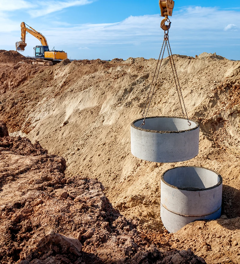 heavy machine hook placing sewer rings into place on job site, excavator in distance